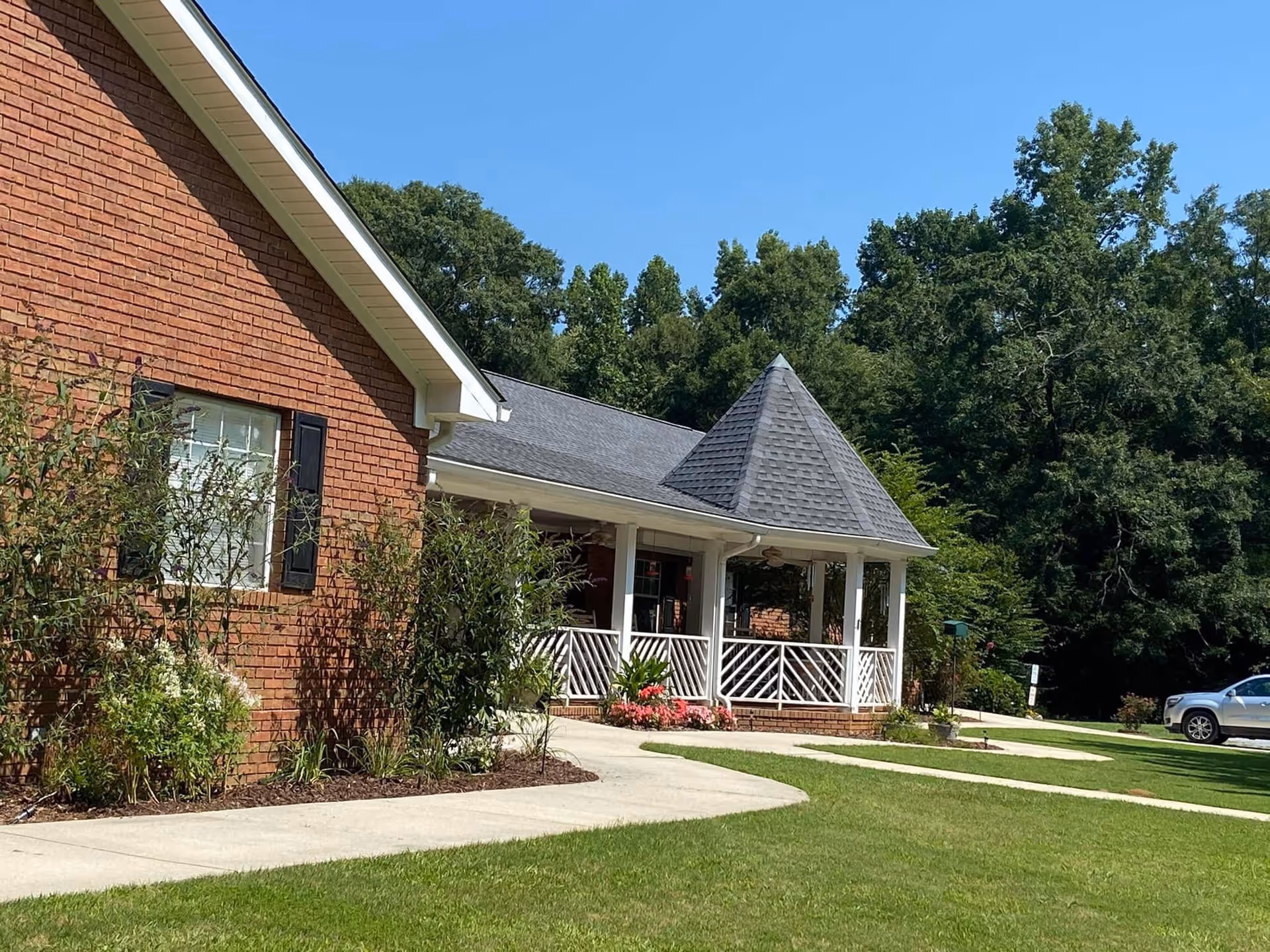 Exterior view of a brick building with a covered porch featuring white railings and a pointed roof section. There are shrubs and flowers planted along the building, a concrete walkway, a grassy lawn, and trees in the background under a clear blue sky.
