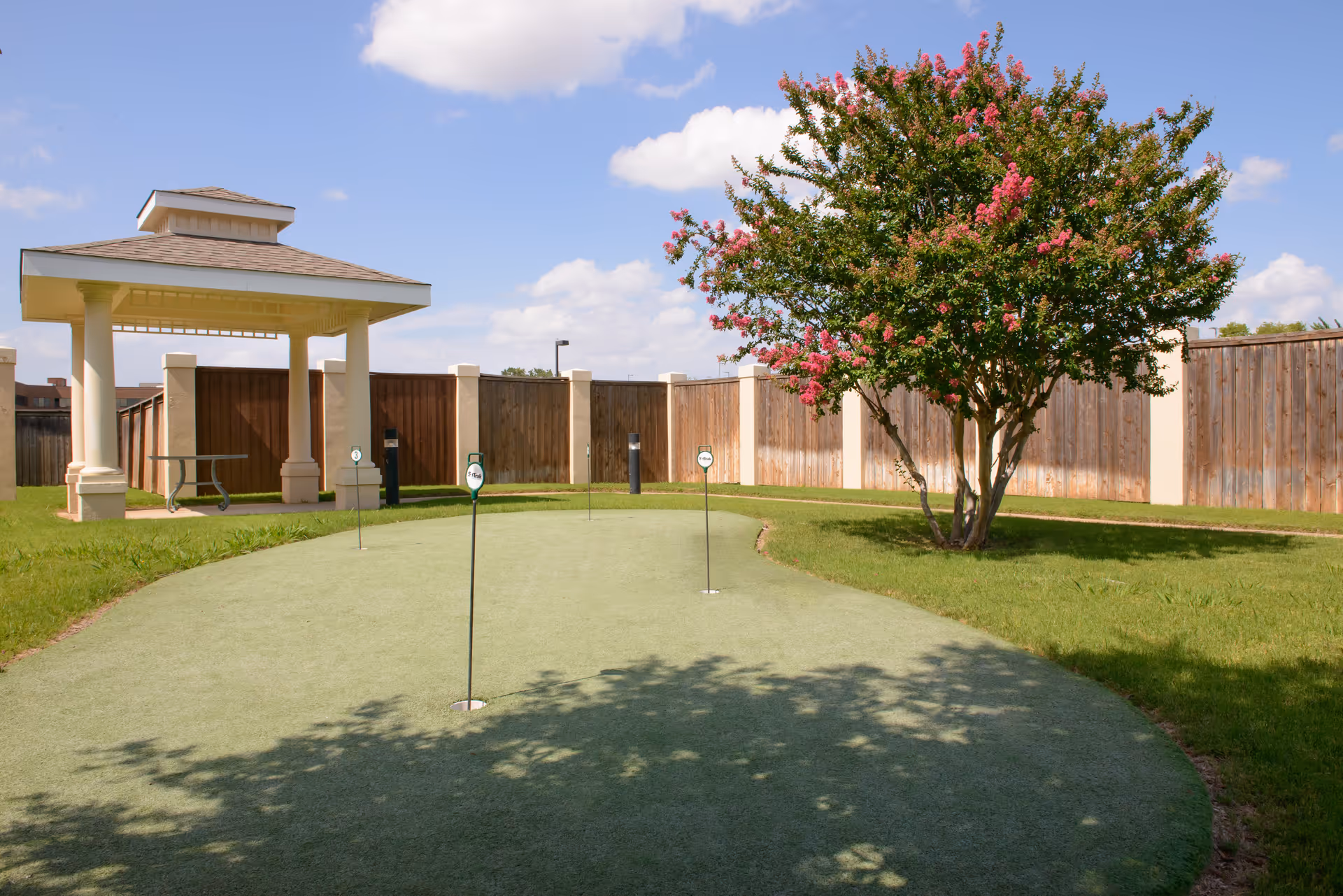 Outdoor putting green with four golf holes and flags, a flowering tree with pink blossoms, a wooden fence surrounding the area, and a small covered pavilion with a picnic table under a blue sky with scattered clouds.