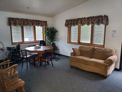 A common area in a senior living facility with a beige couch, a round wooden table surrounded by four chairs, a wheelchair, two wooden rocking chairs, a potted plant, and windows with floral valances.
