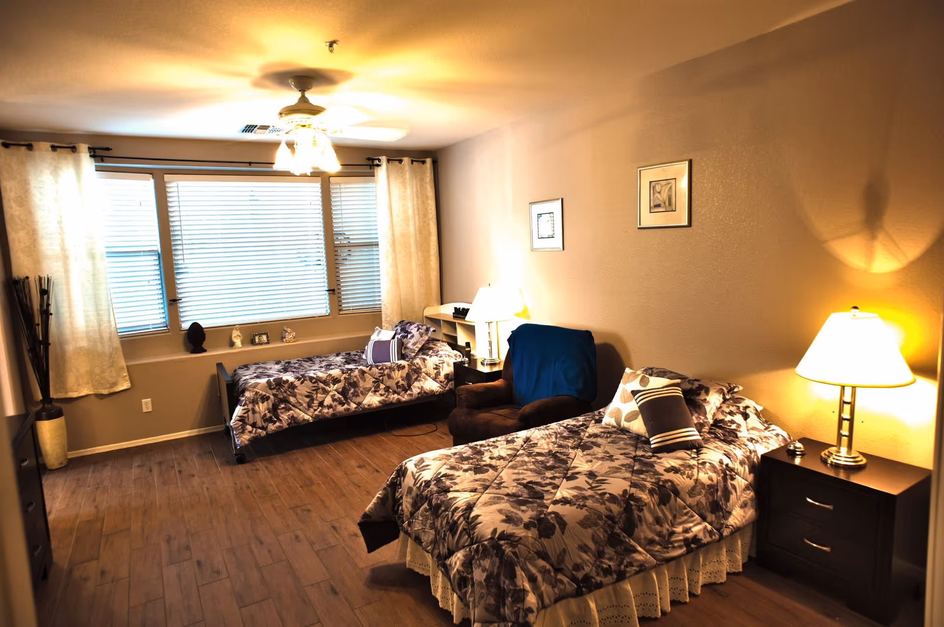 Two twin beds, a chair, nightstands, lamps, and a large window in a warmly lit bedroom.