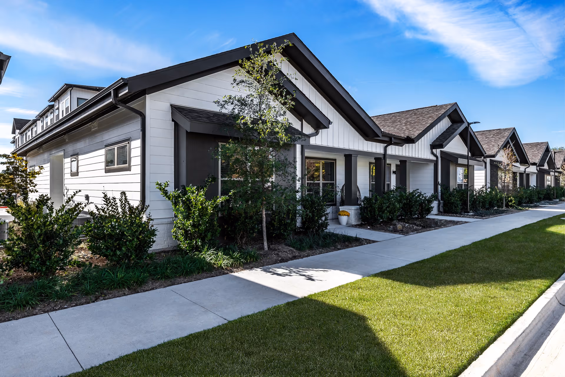 Row of single-story white cottages with porches, shrubs, and a sidewalk under a blue sky.