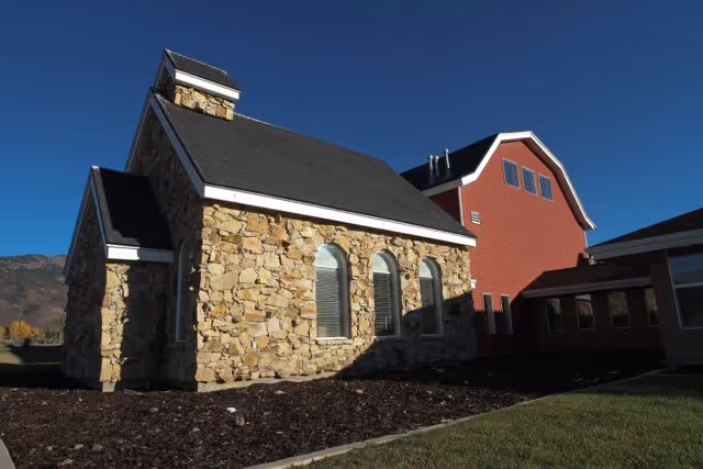 Exterior view of a building at Elk Meadows Assisted Living & Memory Care featuring a stone facade section with three arched windows and a steep dark roof, adjacent to a red barn-style structure with multiple windows under a clear blue sky.