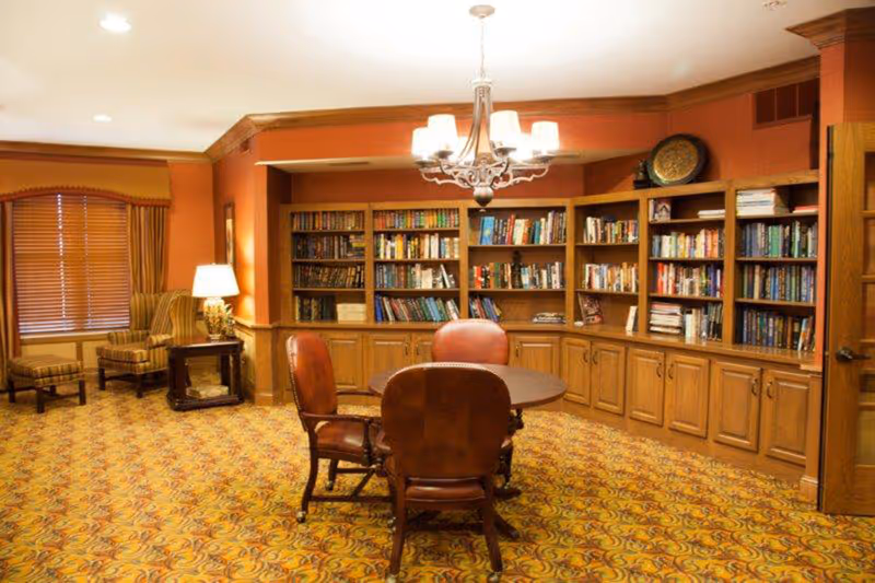 Warm senior living lounge with bookshelves, a round table, and upholstered chairs.