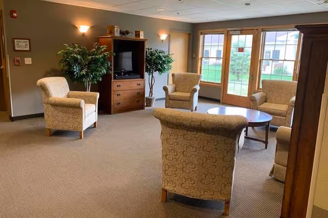A cozy living room area in an assisted living facility featuring four upholstered armchairs arranged around a round wooden coffee table. There is a wooden entertainment center with a TV and two large potted plants on either side. Large windows and a glass door provide natural light and a view of the outdoor greenery.