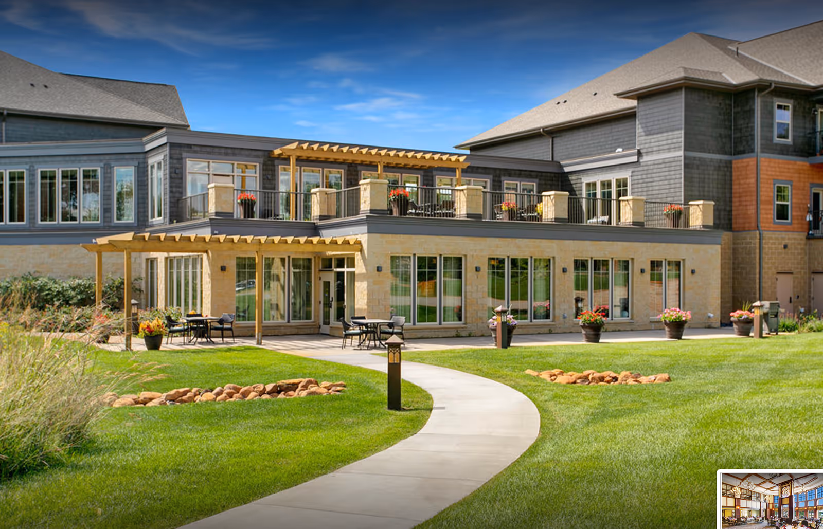 Modern two-story senior living building with patios, balconies, and a curved walkway through a manicured lawn.