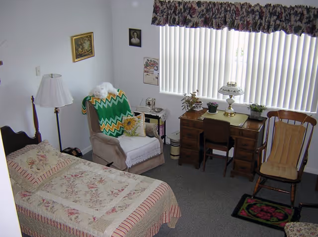 A cozy bedroom with a single bed covered in a floral quilt, a cushioned armchair draped with a green and yellow zigzag blanket, a wooden desk with a lamp and potted plants, a wooden rocking chair with a cushion, and vertical blinds covering the window.