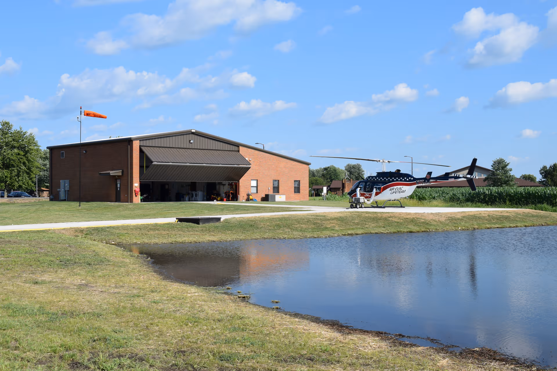A brick facility exterior with a hangar opening and a helicopter parked near a pond.
