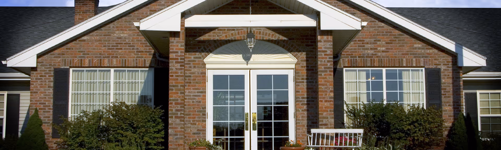 Brick-front entrance with double glass doors, flanking windows, a white bench, and shrubs.
