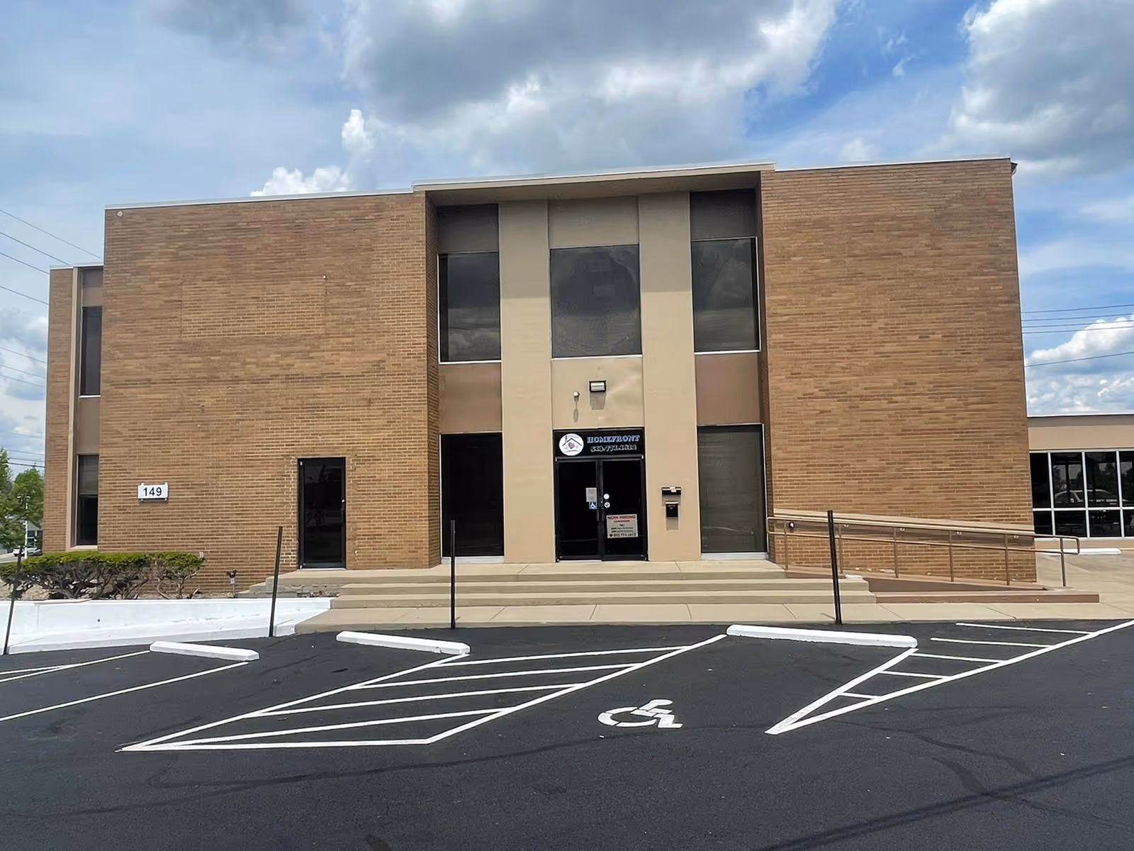Brick building exterior with central entrance and accessible ramp, parking lot in foreground.