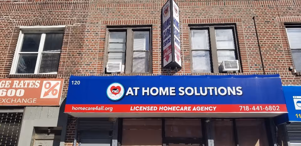 Brick storefront with a blue AT HOME SOLUTIONS sign and licensed homecare banner.