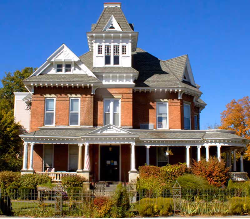 A brick Victorian-style assisted living building with a wraparound porch and manicured garden.