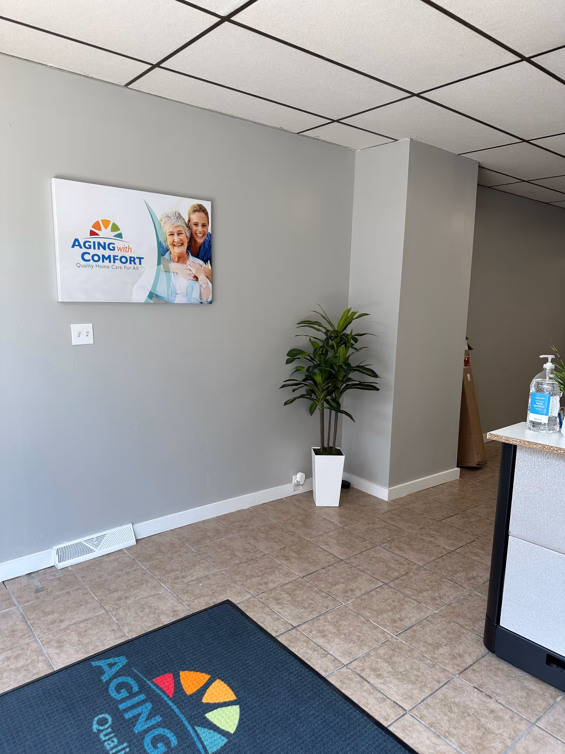 Interior lobby area with Aging with Comfort sign, potted plant, and sanitizer on a counter.