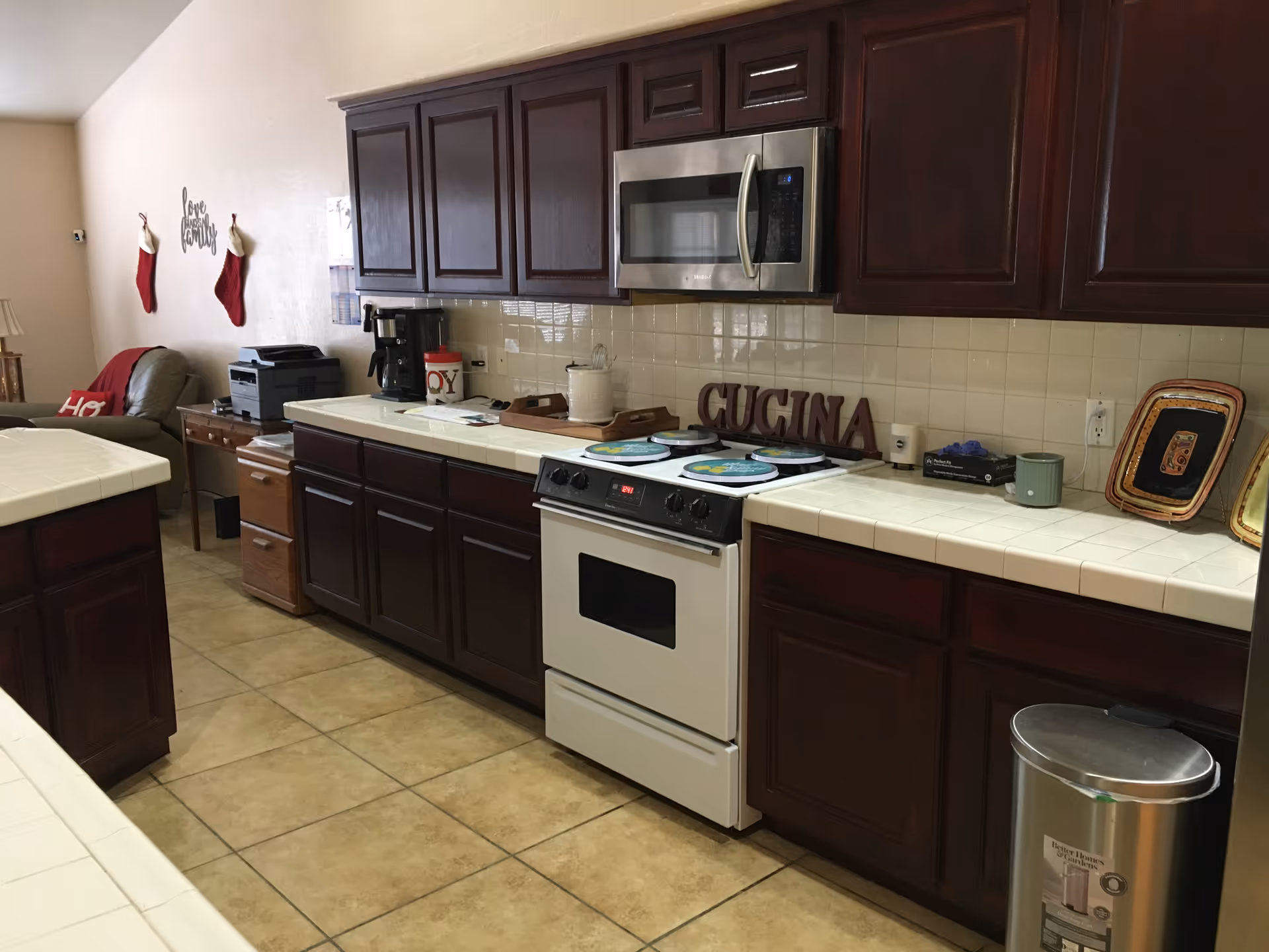 Long kitchen with dark wood cabinets, white appliances, and tiled counters in a senior living facility.