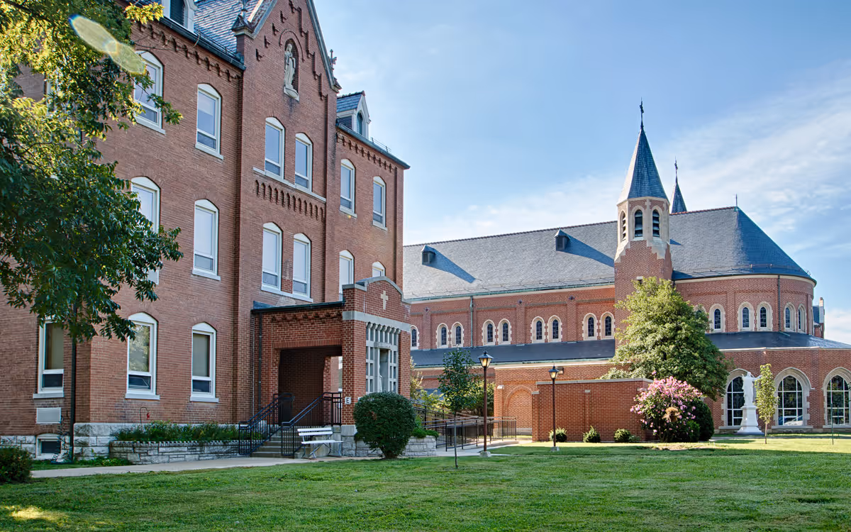 Front view of a red-brick senior living building and attached chapel-like structure with a lawn and trees under a blue sky.