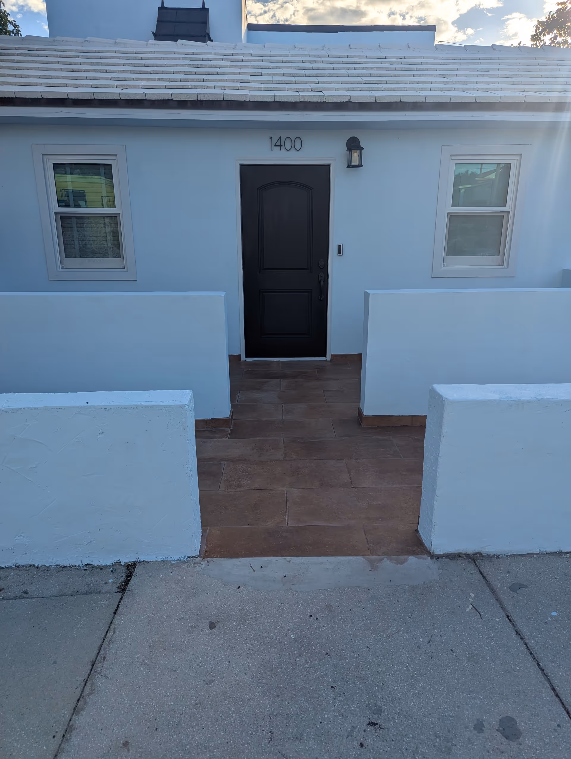 White building entrance with a black door, two windows, and a tiled walkway.