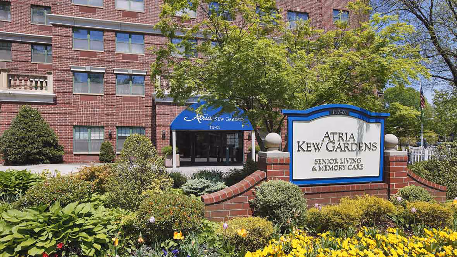 Exterior view of Atria Kew Gardens senior living and memory care facility showing a brick building with multiple windows, a blue awning over the entrance, and a landscaped garden with various green plants and yellow flowers in front of a sign displaying the facility's name.