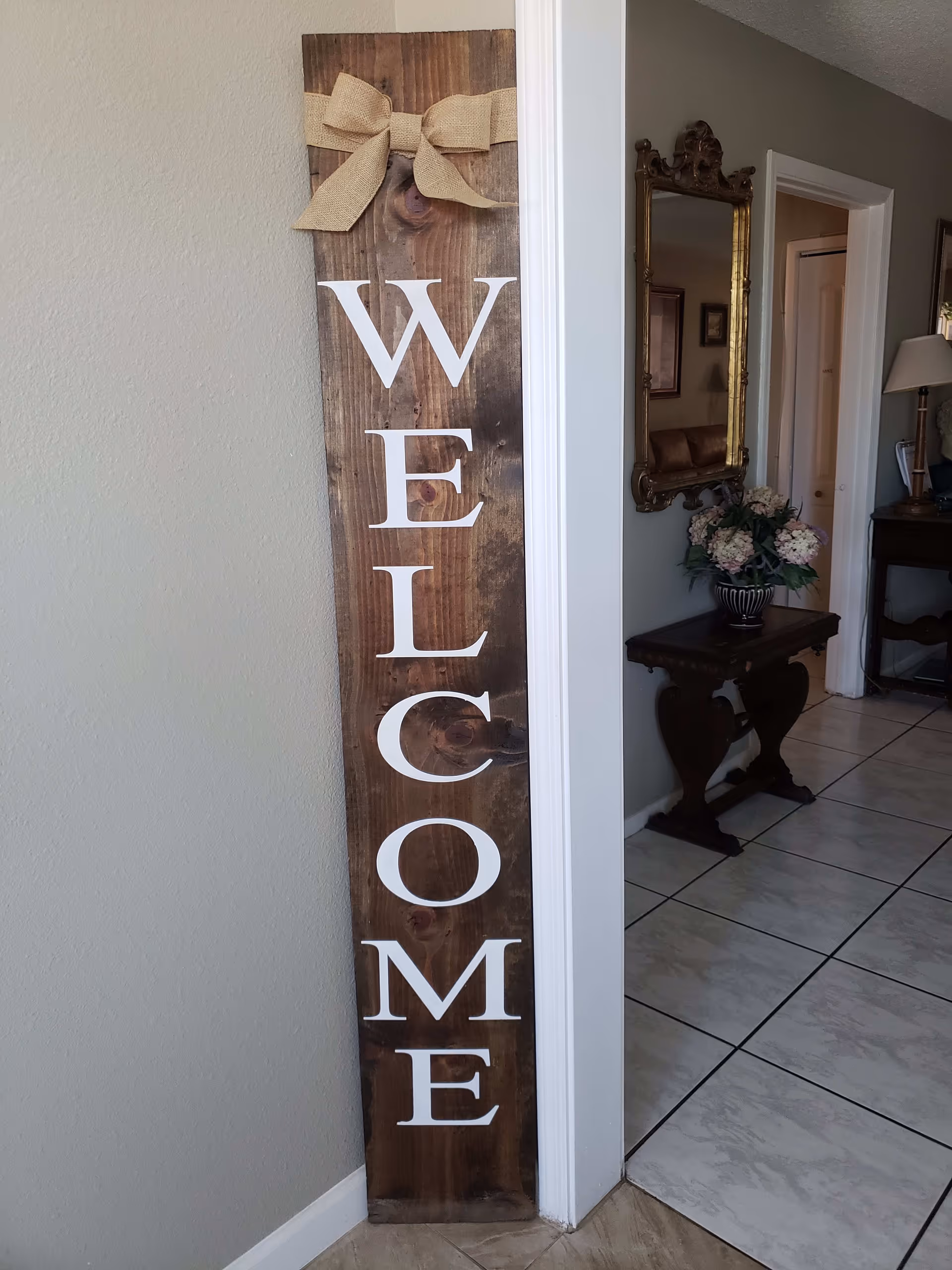 A tall wooden 'WELCOME' sign leans against an interior wall beside a hallway with a mirror, side table and flower arrangement.