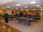 A spacious dining room with multiple tables and blue chairs arranged neatly. The room has beige carpeting, wooden paneling on the walls, and several large potted plants. The ceiling has recessed lighting, and there are framed pictures on the walls.