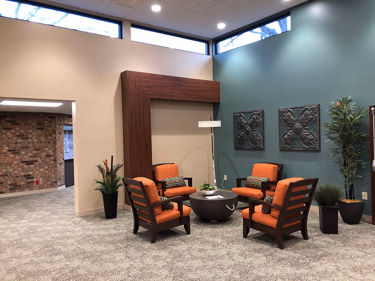 A cozy seating area with four wooden chairs featuring orange cushions arranged around a round black coffee table. The room has a teal accent wall with two decorative metal wall hangings and several potted plants. There is a tall floor lamp next to the chairs and a large wooden frame on the beige wall. The floor is carpeted with a patterned design, and there are high windows near the ceiling letting in natural light.