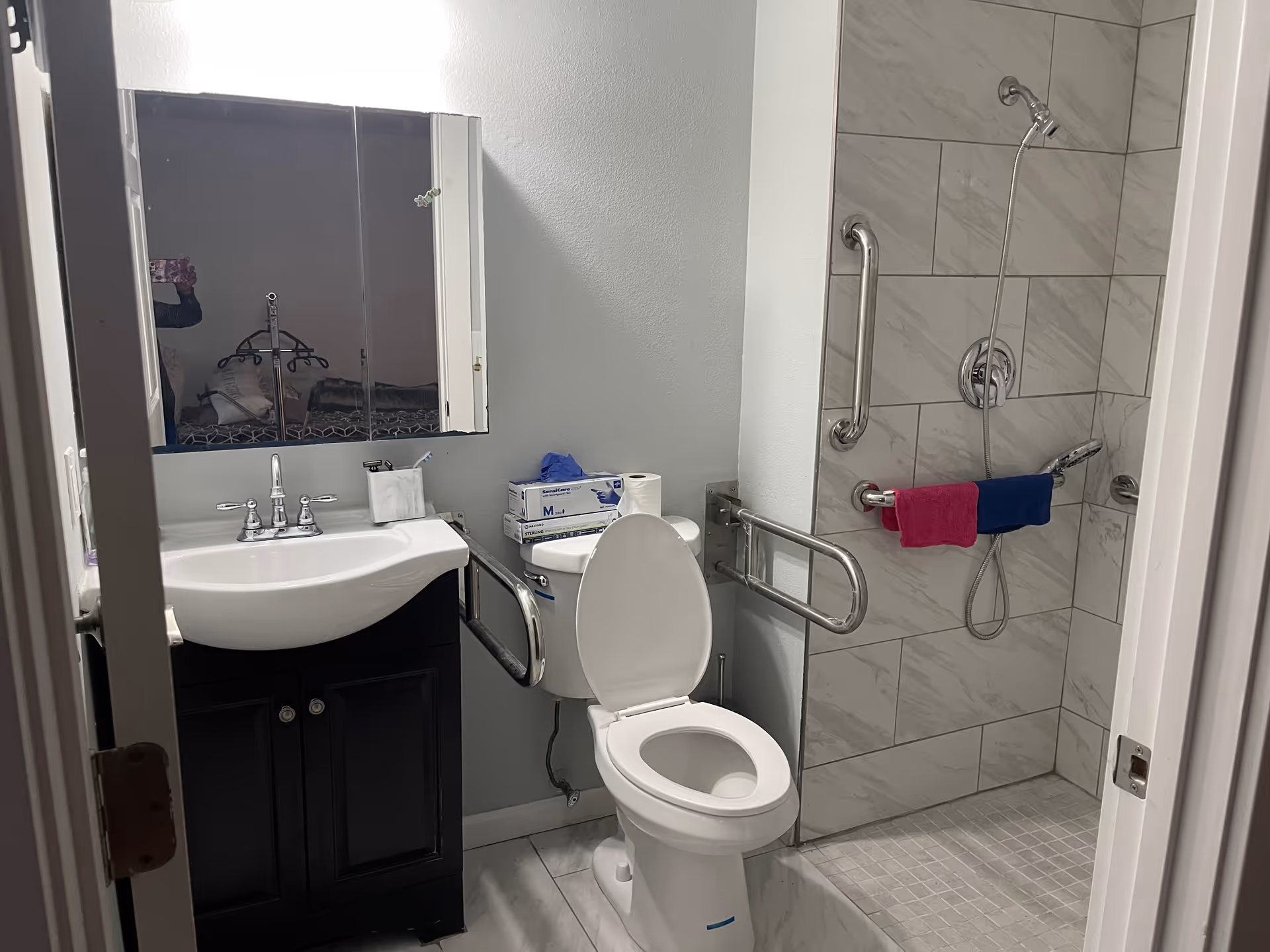 Accessible bathroom with a sink and mirrored medicine cabinet, a toilet with fold-down grab bars, and a tiled walk-in shower with grab bars and towels.
