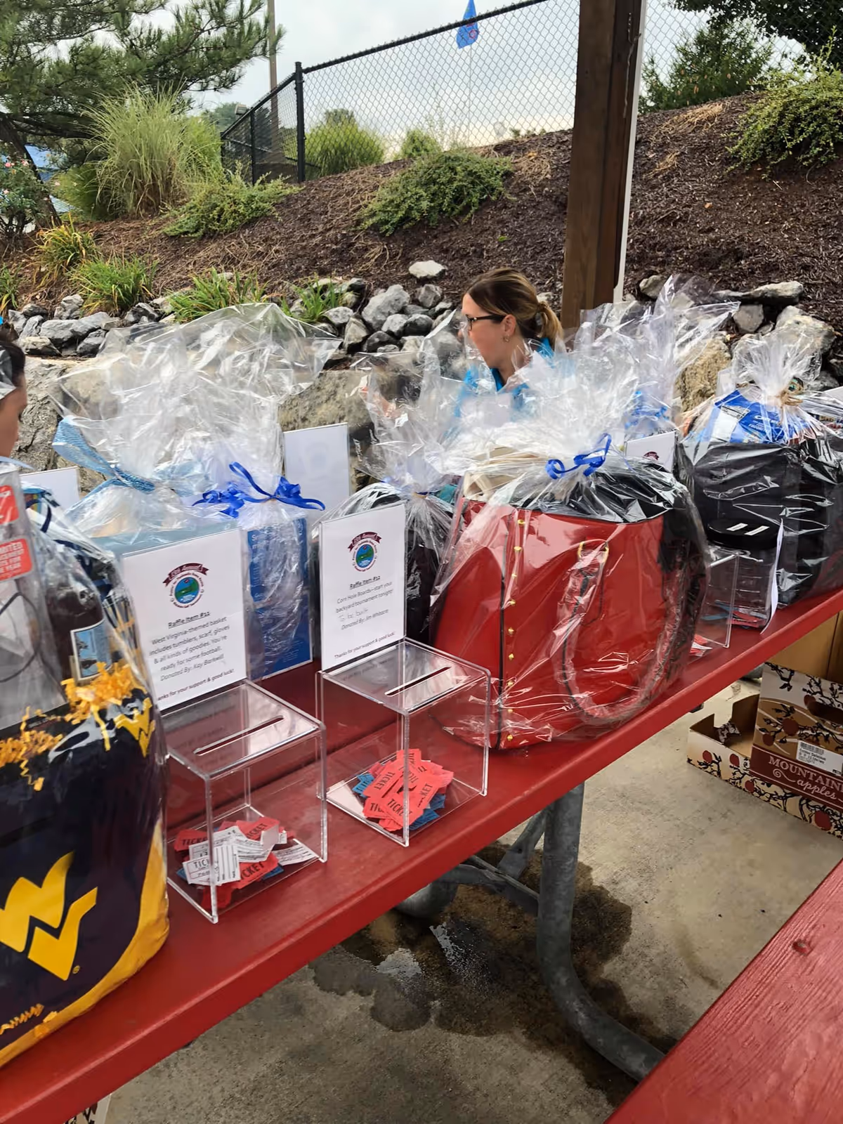 Outdoor charity event with gift baskets and a woman arranging items on a red picnic table.