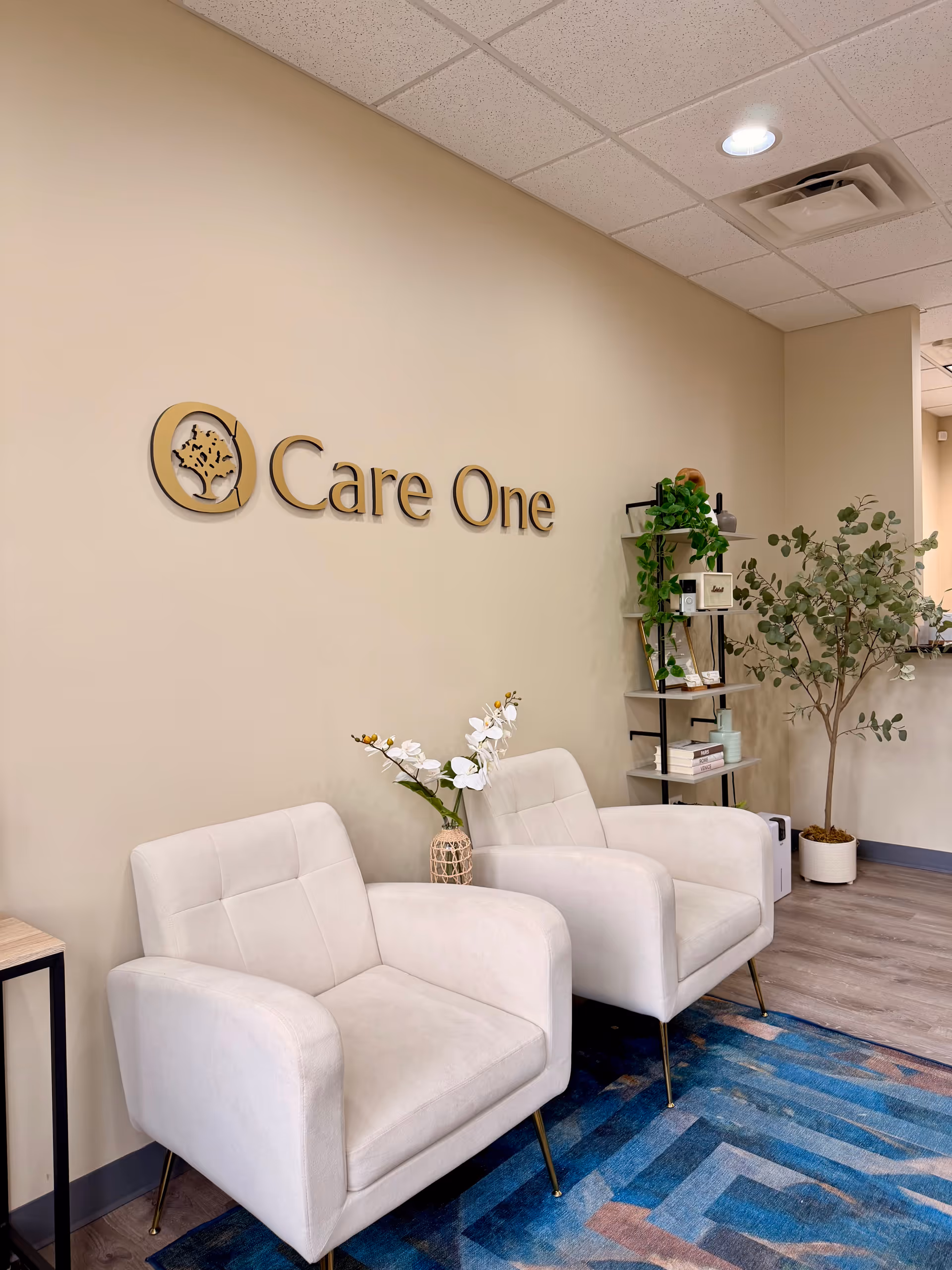 Care One lobby with two white armchairs, plants, and a beige wall sign.