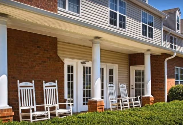 Front porch of a brick senior living building with white rocking chairs.