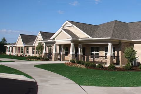 Exterior view of a single-story senior living facility building with multiple entrances, beige brick walls, white trim, and a gray shingled roof under a clear blue sky. The building is surrounded by well-maintained green lawns, small shrubs, and a concrete walkway.
