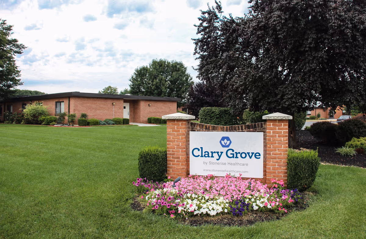 A landscaped outdoor area with a brick sign that reads 'Clary Grove by Stonerise Healthcare' surrounded by colorful flowers and green grass, with a single-story brick building and trees in the background under a partly cloudy sky.