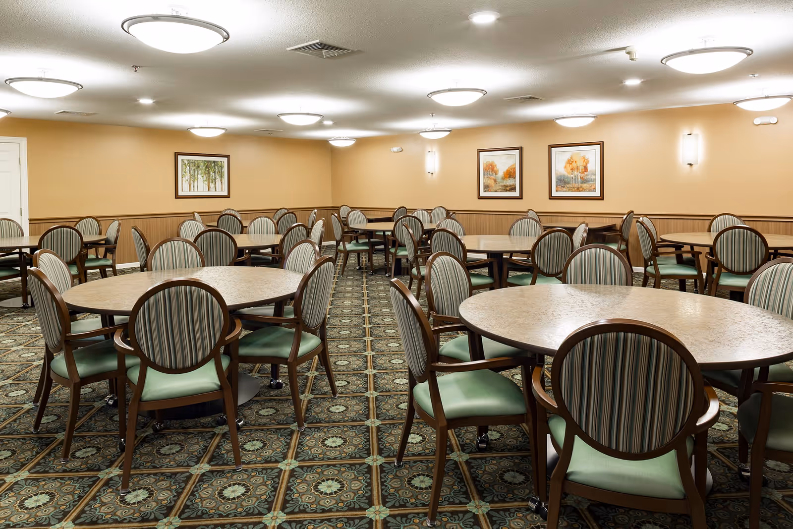 A spacious dining room with multiple round tables surrounded by wooden chairs with green cushioned seats and striped backs. The room has beige walls adorned with framed artwork and is well-lit by ceiling lights and wall sconces. The floor is covered with a patterned carpet.