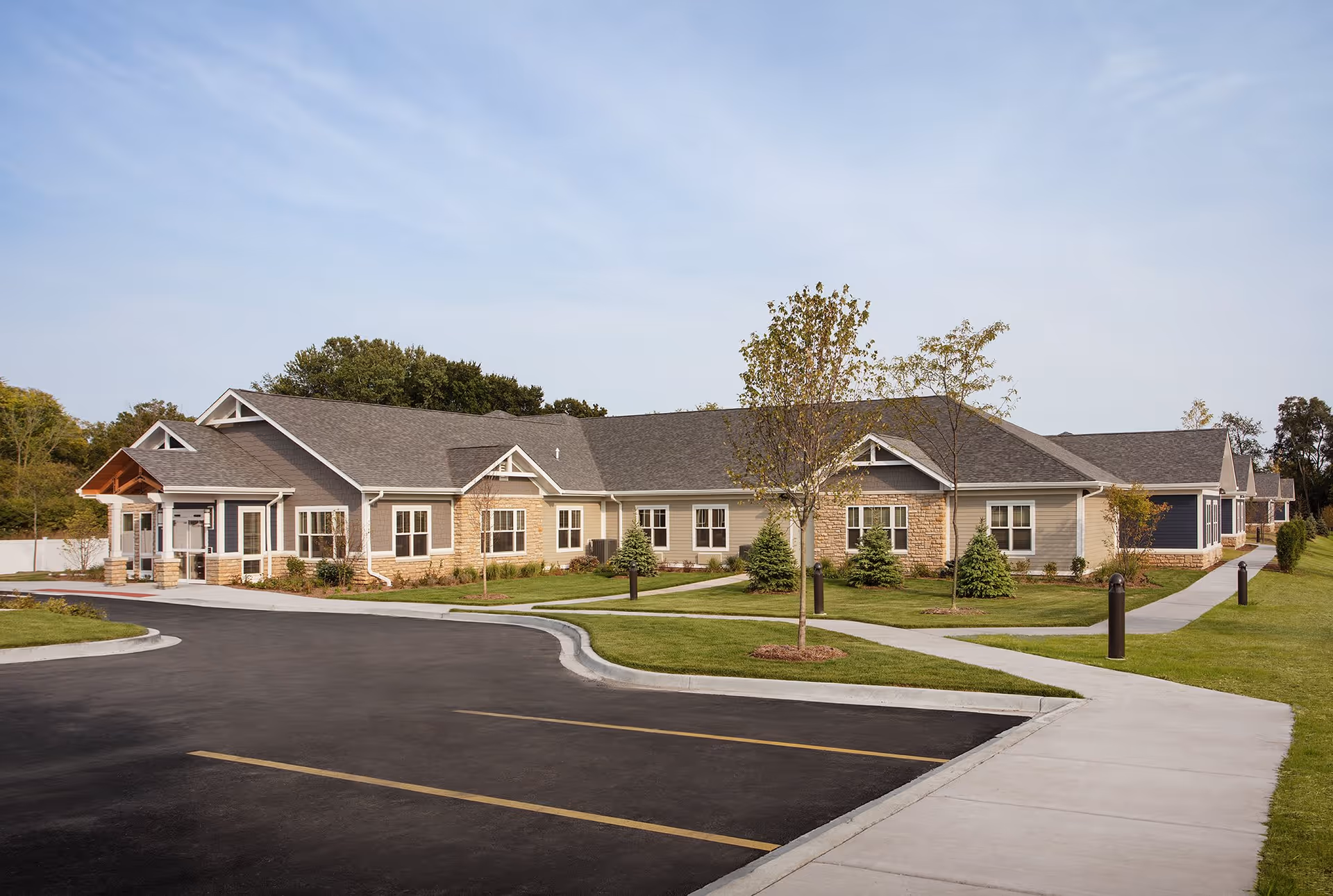 Exterior view of a single-story senior living facility building with beige and gray siding, stone accents, multiple windows, and a peaked roof. The building is surrounded by a well-maintained lawn with young trees and shrubs, a paved parking lot, and sidewalks leading to the entrance.