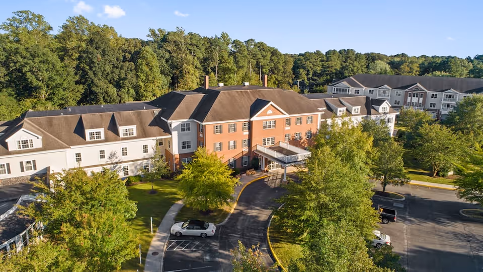 Aerial view of Lakeside At Mallard Landing senior living facility showing a large multi-story building with a mix of brick and white siding exterior surrounded by trees and greenery, with a parking lot in front and a clear blue sky above.