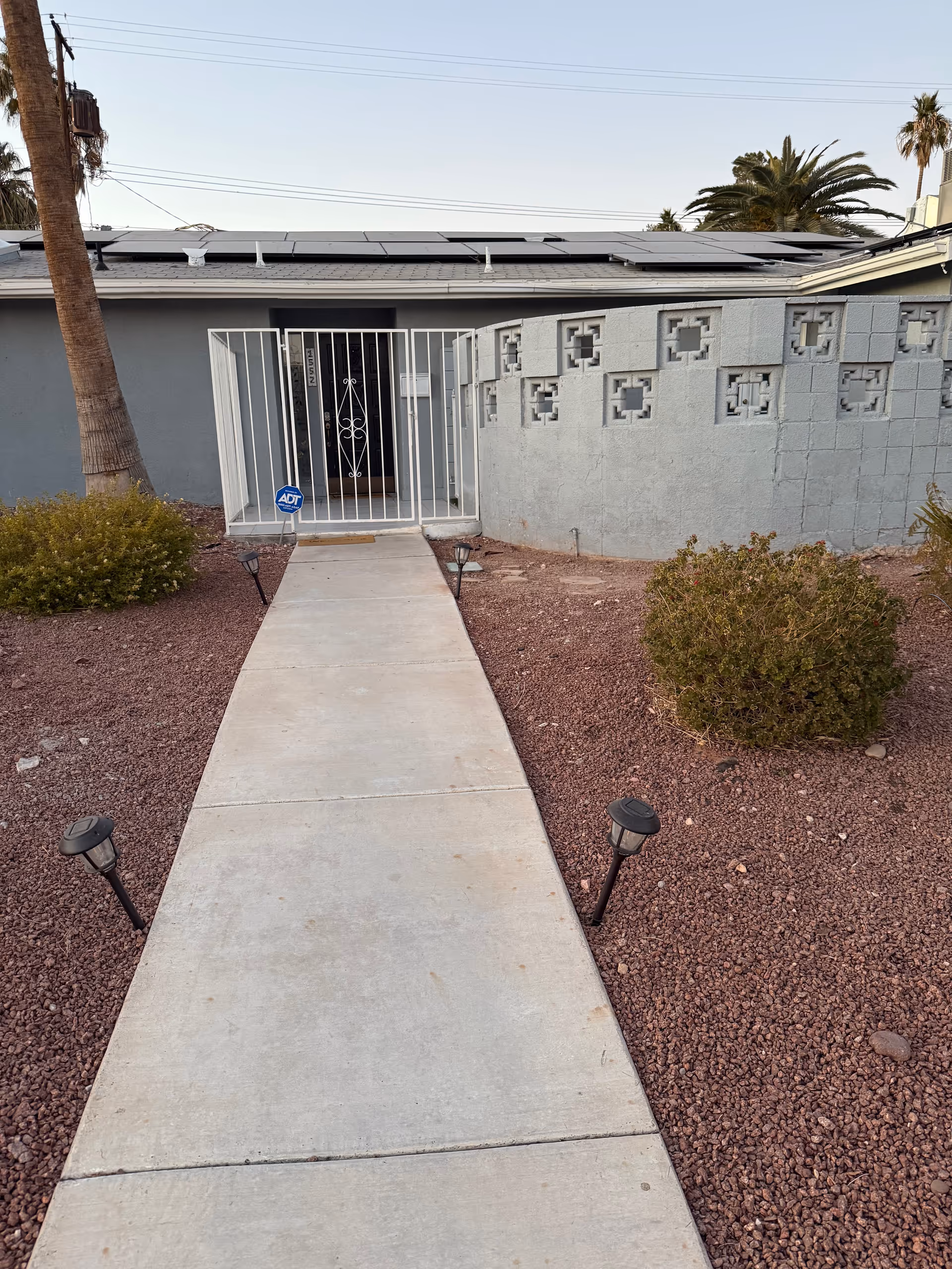 Concrete walkway leads to a metal gate entrance of a single-story building with desert landscaping.