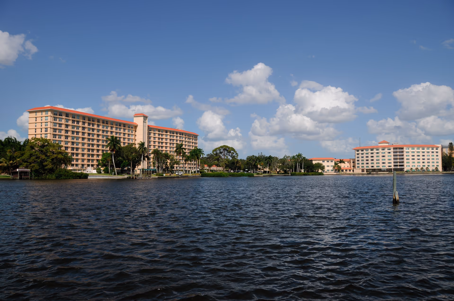View of two large multi-story buildings with red roofs situated along a waterfront under a blue sky with scattered clouds. Palm trees and greenery are visible around the buildings.