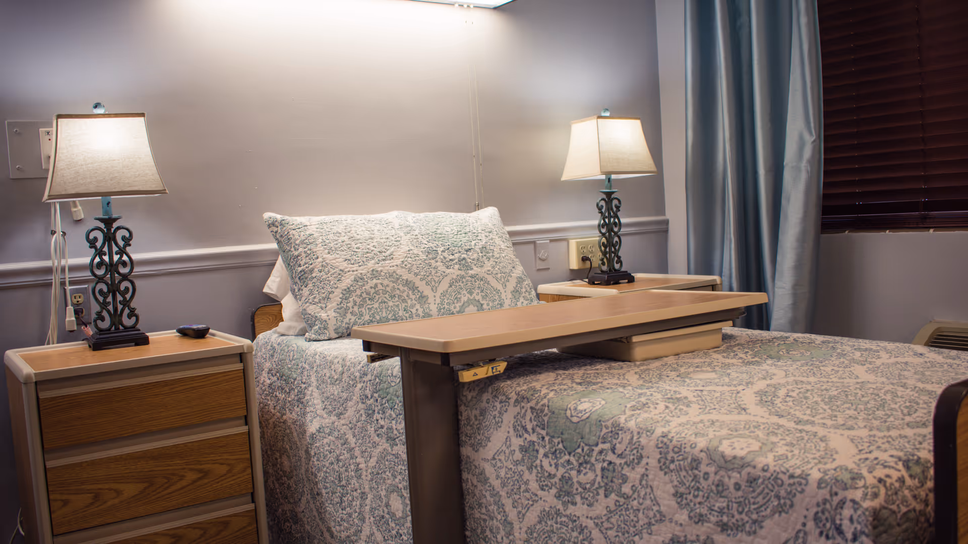 A neatly made bed with a patterned bedspread and pillow in a healthcare facility room. There are two bedside tables, each with a decorative lamp. A wooden overbed table is positioned over the bed. The room has light-colored walls, a window with closed blinds, and blue curtains.