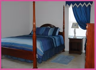 A bedroom with a four-poster bed dressed in blue linens, a nightstand and lamp, blue curtains, and a small rug on a tiled floor.