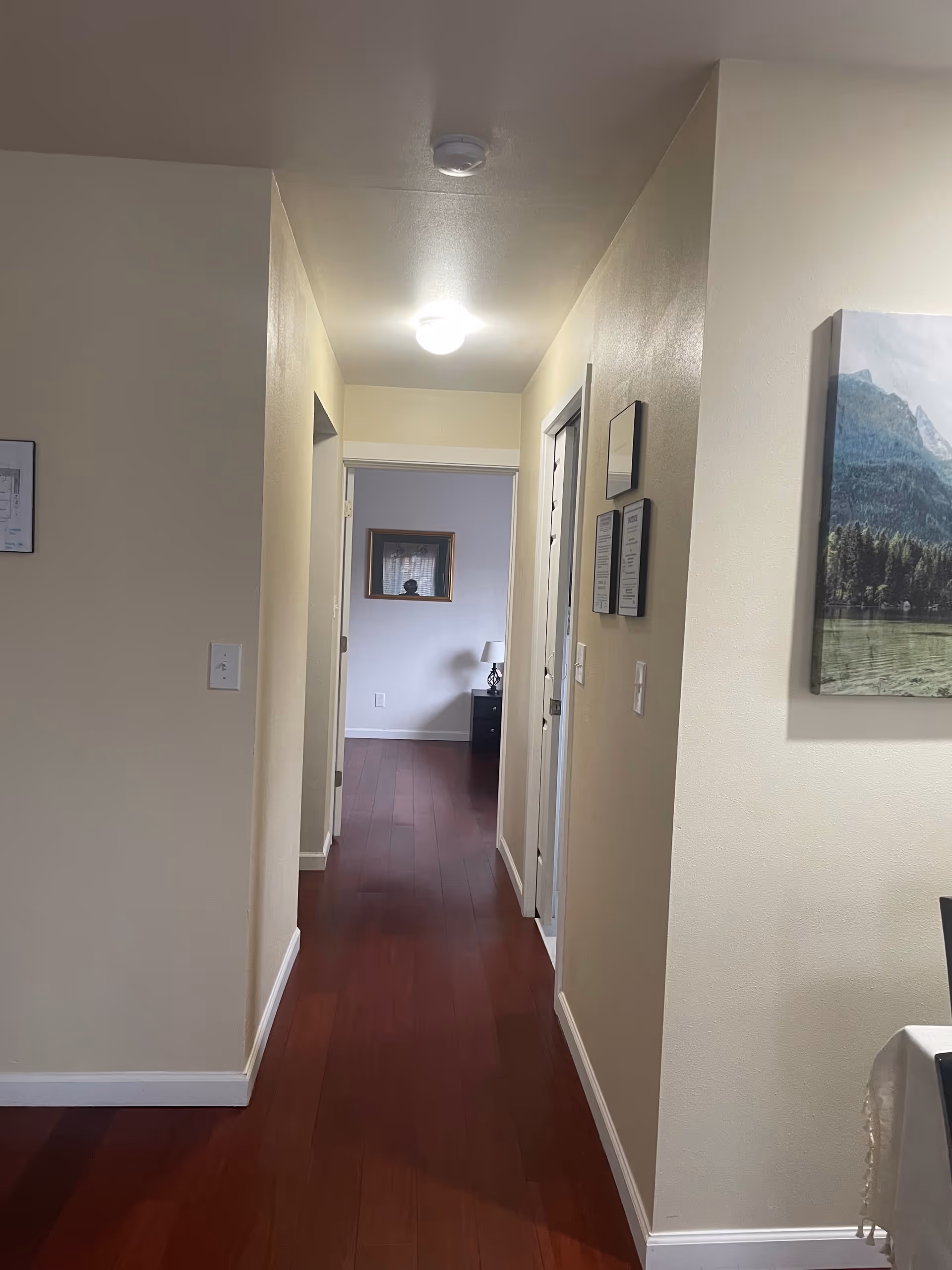 A narrow corridor in a senior living facility with beige walls, hardwood floor, and framed pictures.
