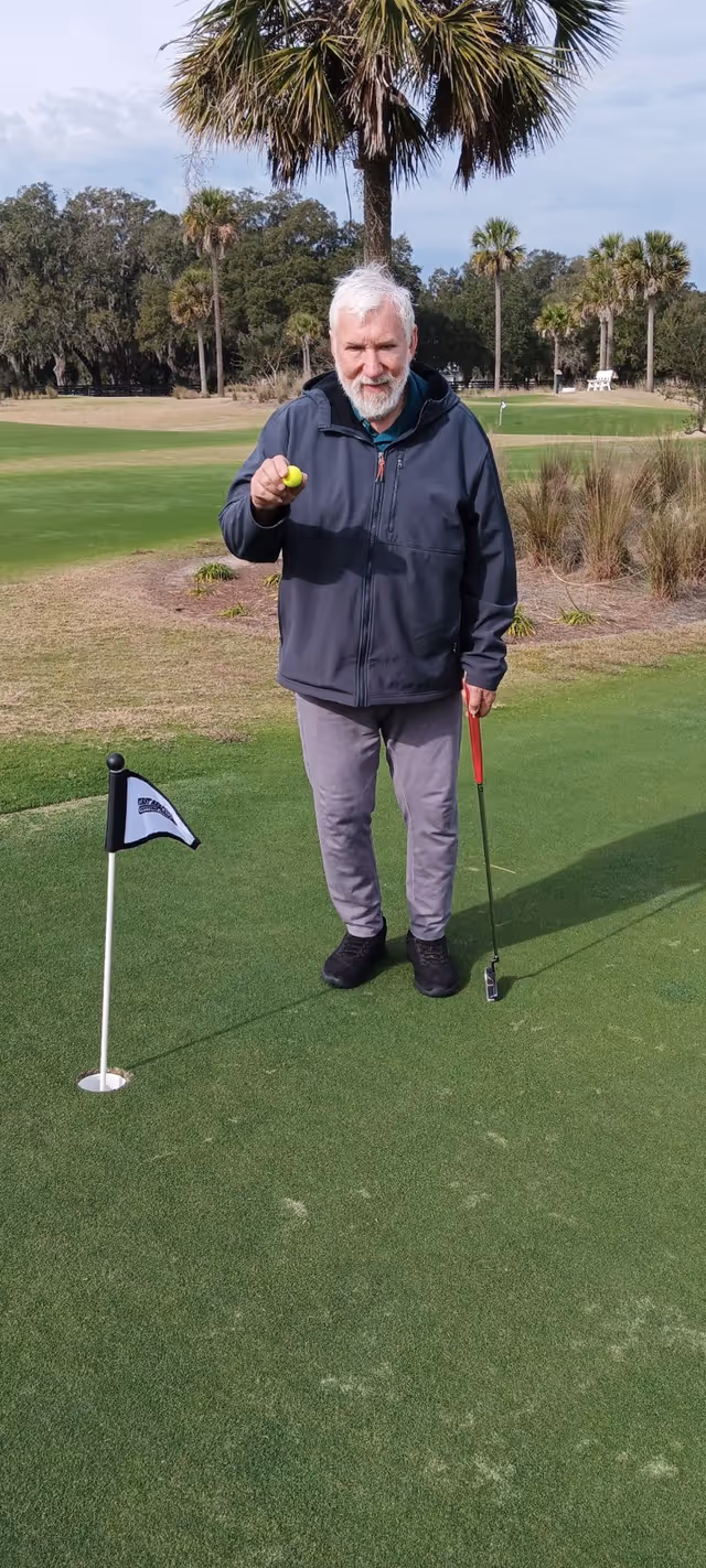 Older man on a golf putting green holds a ball beside a flag on sunny course.