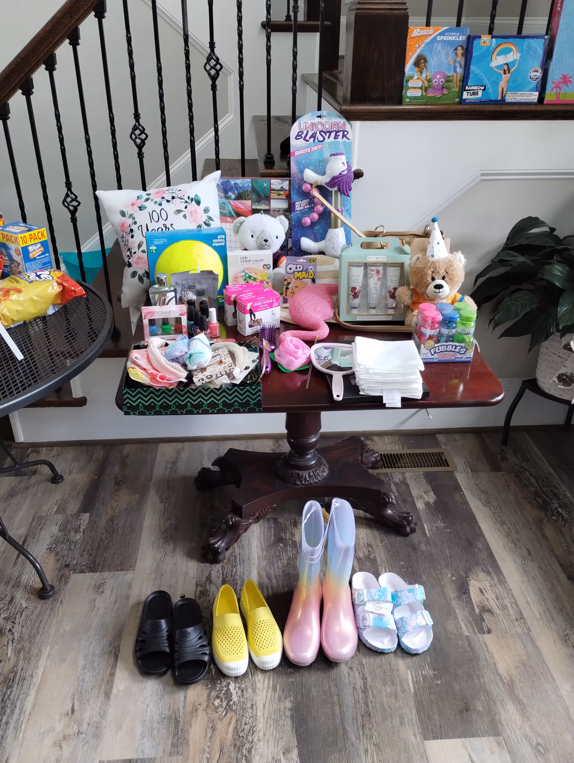 A wooden table in a home interior setting with various items displayed on top, including stuffed animals, a unicorn blaster toy, nail polish, skincare products, bubbles, a pillow with floral design, and other small gift items. In front of the table on the floor are four pairs of shoes: black slip-ons, yellow slip-ons, pastel-colored rain boots, and white sandals. The background shows a staircase with black iron railings and a potted plant to the right.