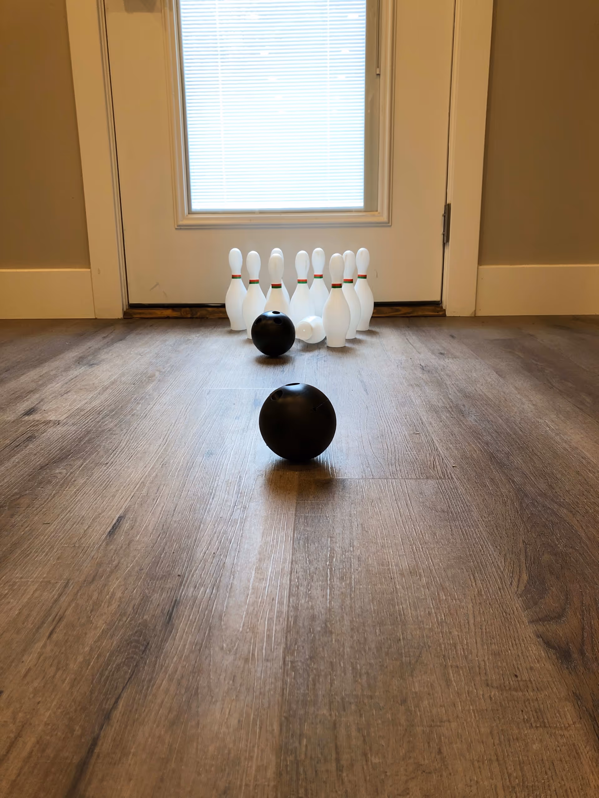 Small indoor bowling setup with plastic pins and two black mini bowling balls arranged on a wood floor in front of a door.