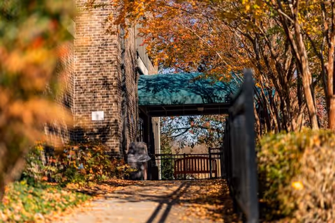 Autumn leaves line a brick walkway toward the facility entrance under a green canopy.