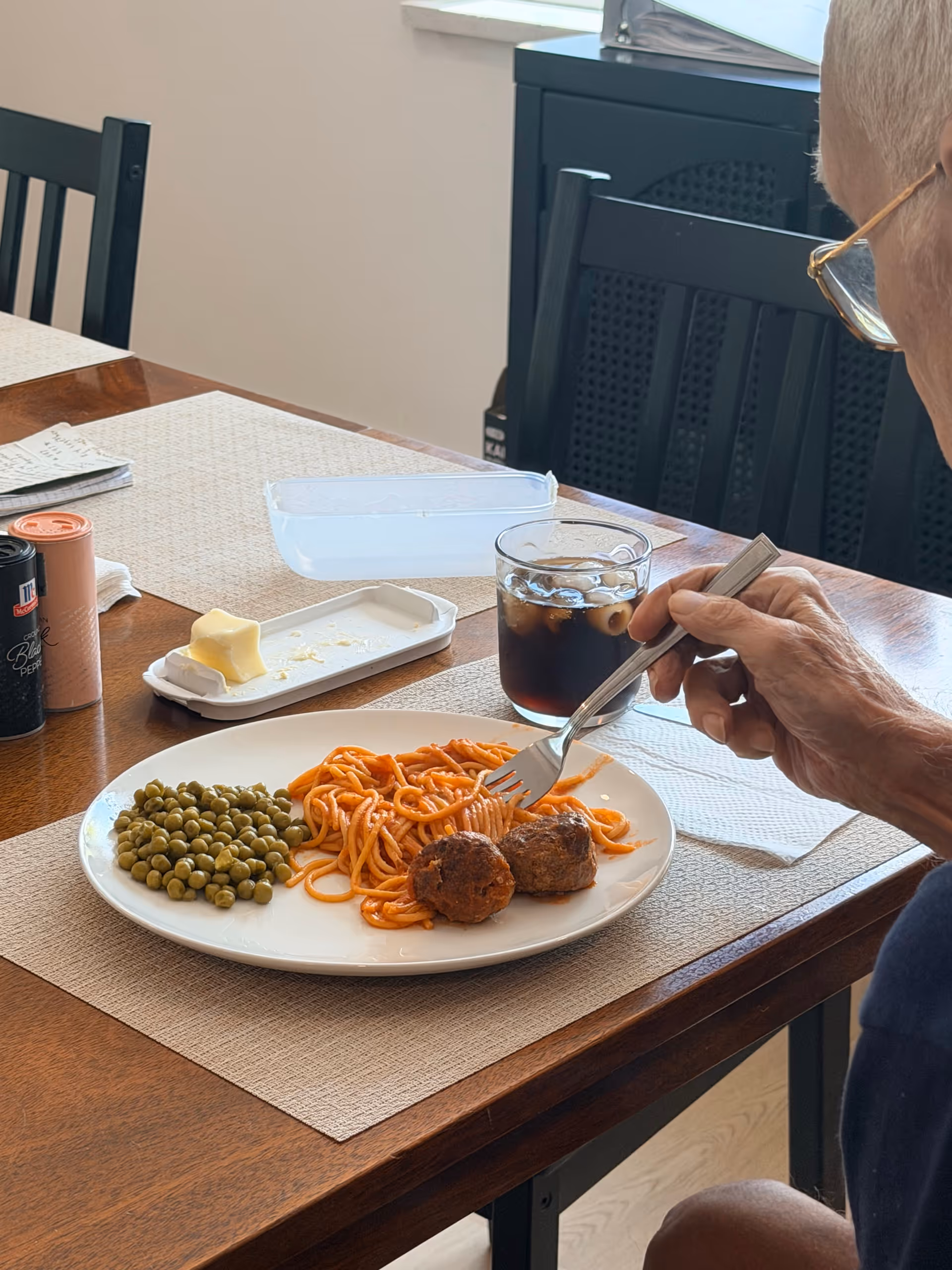 An elderly person eats spaghetti with meatballs and peas at a dining table.