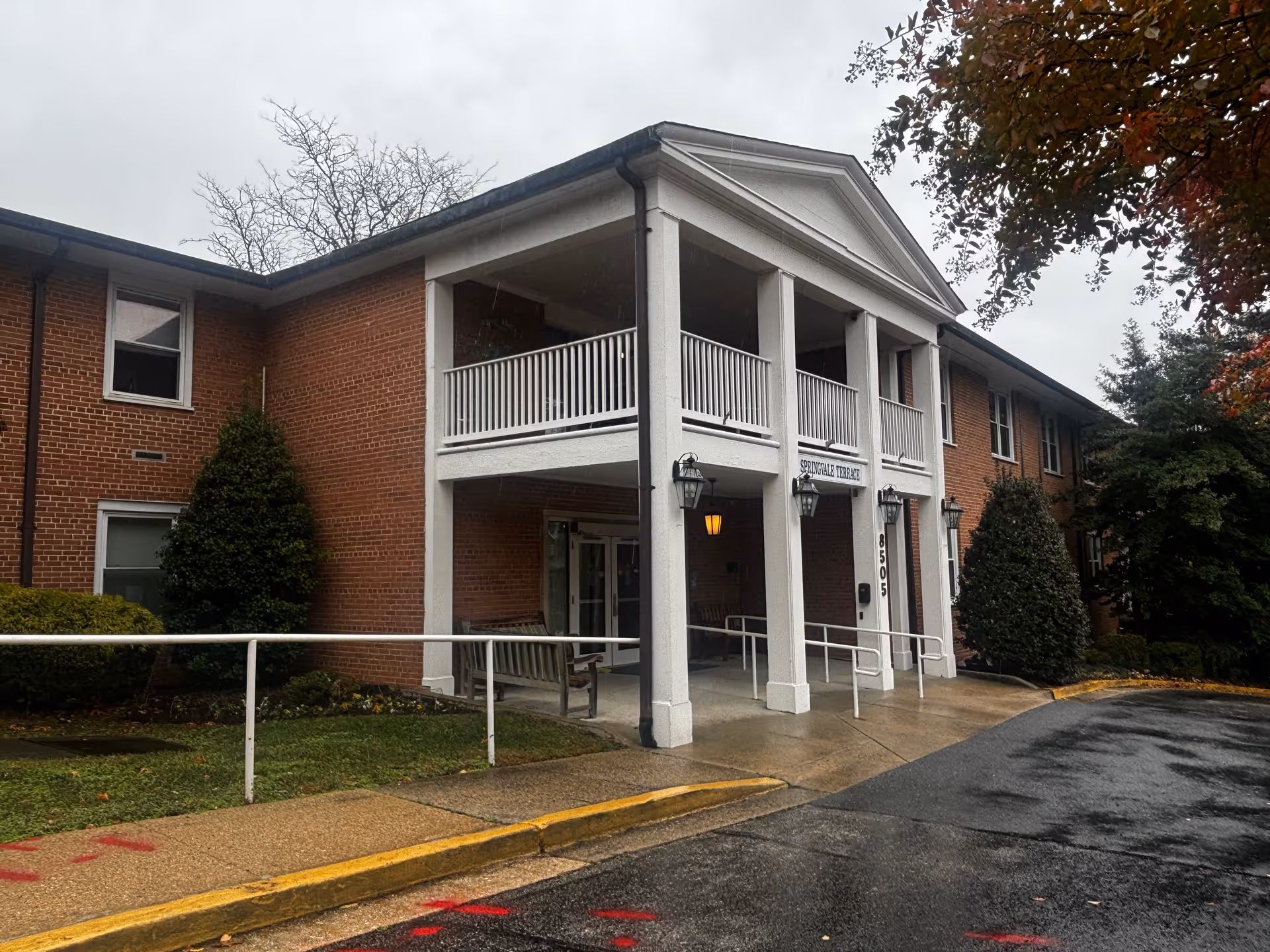 Exterior view of a two-story brick building with white columns and a covered entrance. The building has a sign that reads 'Springvale Terrace' and the address number 8505 is visible on a column. There are bushes and trees around the building, and the ground is wet from rain.