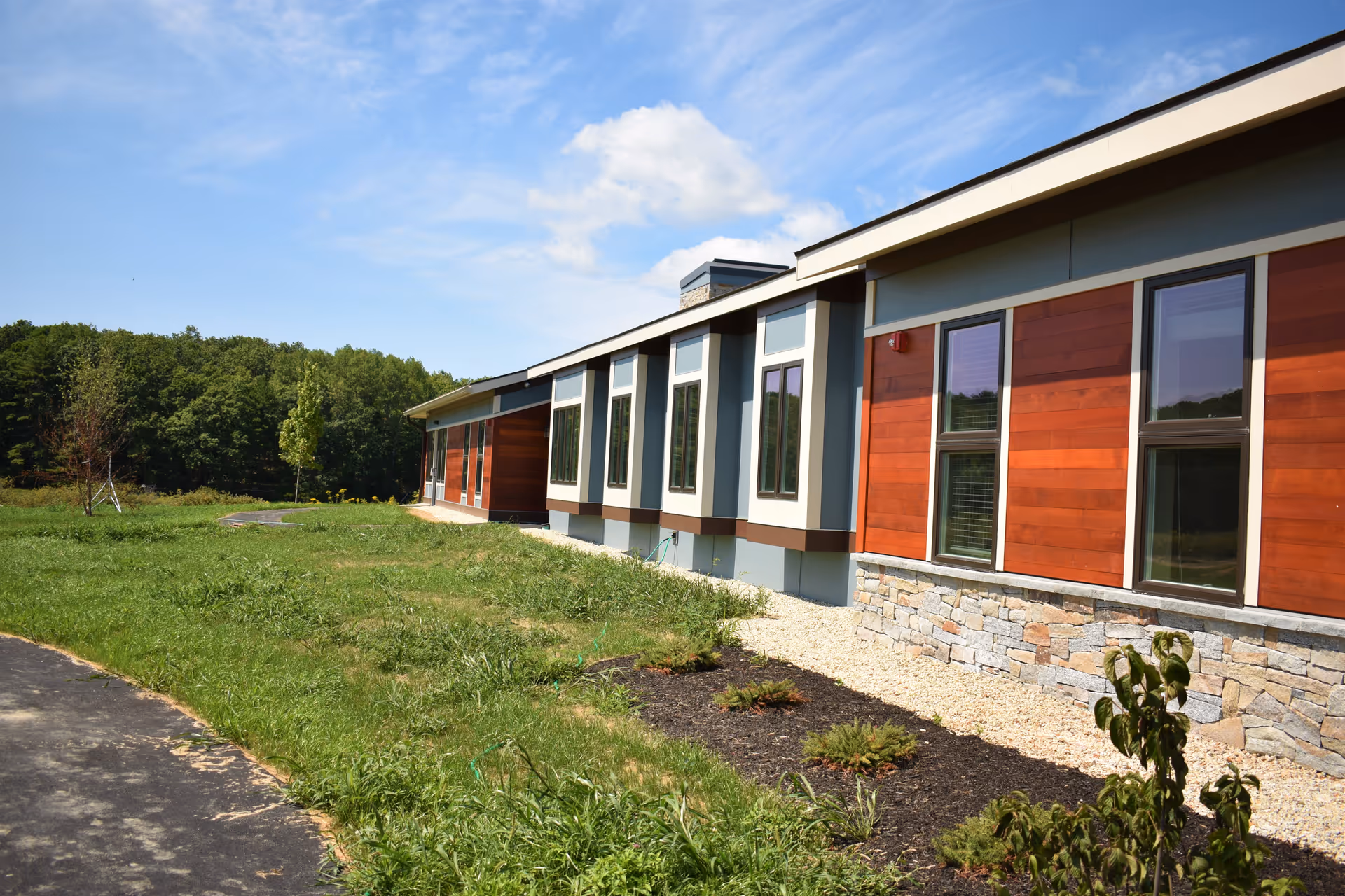 Exterior view of a modern single-story building with large windows, wood paneling, stone accents, and a paved walkway surrounded by grass and small plants under a partly cloudy blue sky.