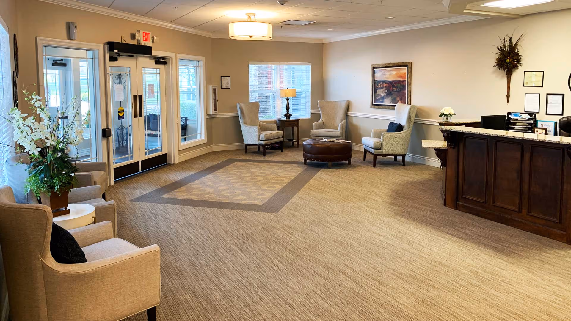A spacious and well-lit reception area with beige walls and carpet. There are several comfortable armchairs arranged around a round ottoman near a window with blinds. A wooden reception desk is on the right side with some office supplies and framed certificates on the wall behind it. A large floral arrangement is placed on a small round table near the entrance doors, which have glass panels and an exit sign above them.