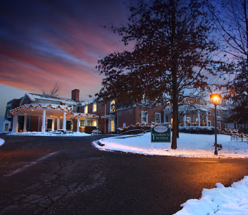 Snow-covered brick building entrance at dusk decorated with holiday lights and a 'Garden Suites' sign.