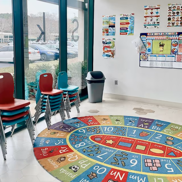 Bright indoor activity room with stacked chairs, an alphabet rug, and posters.