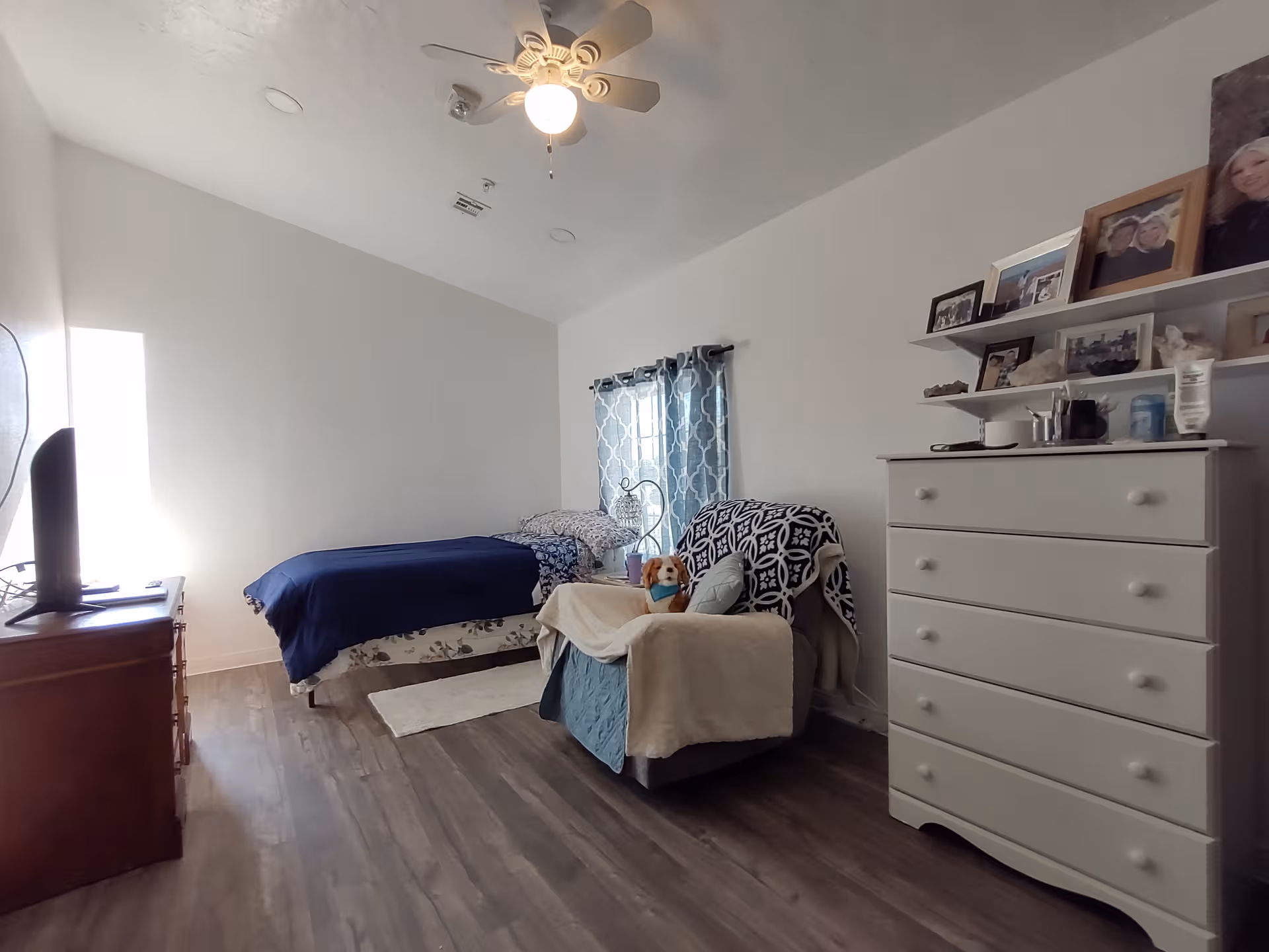 A senior living bedroom with bed, chair, dresser, framed photos, and a window.