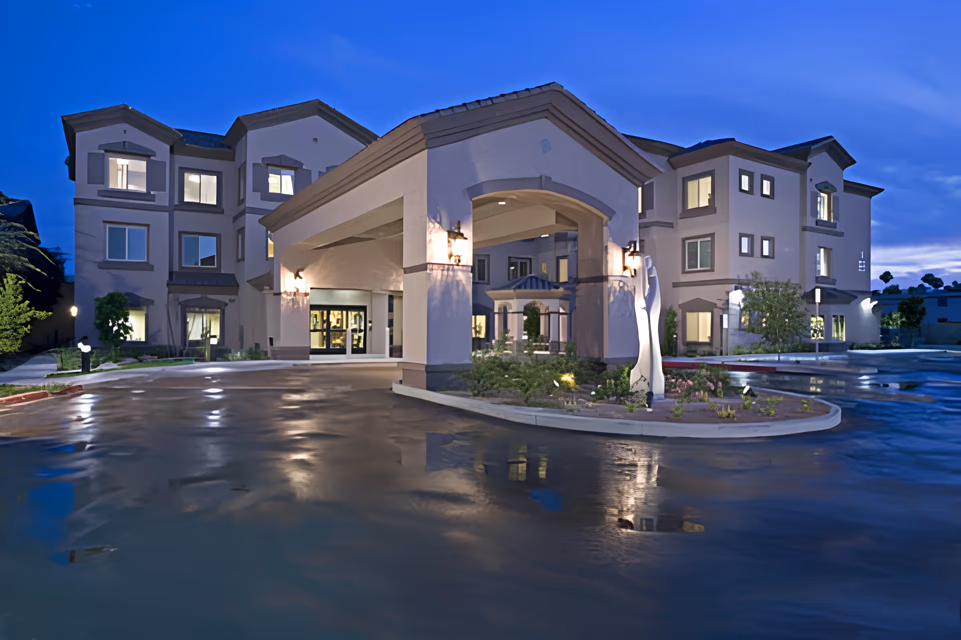 Front entrance of a three-story senior living building with a covered porte-cochere lit at dusk and wet driveway reflecting lights.