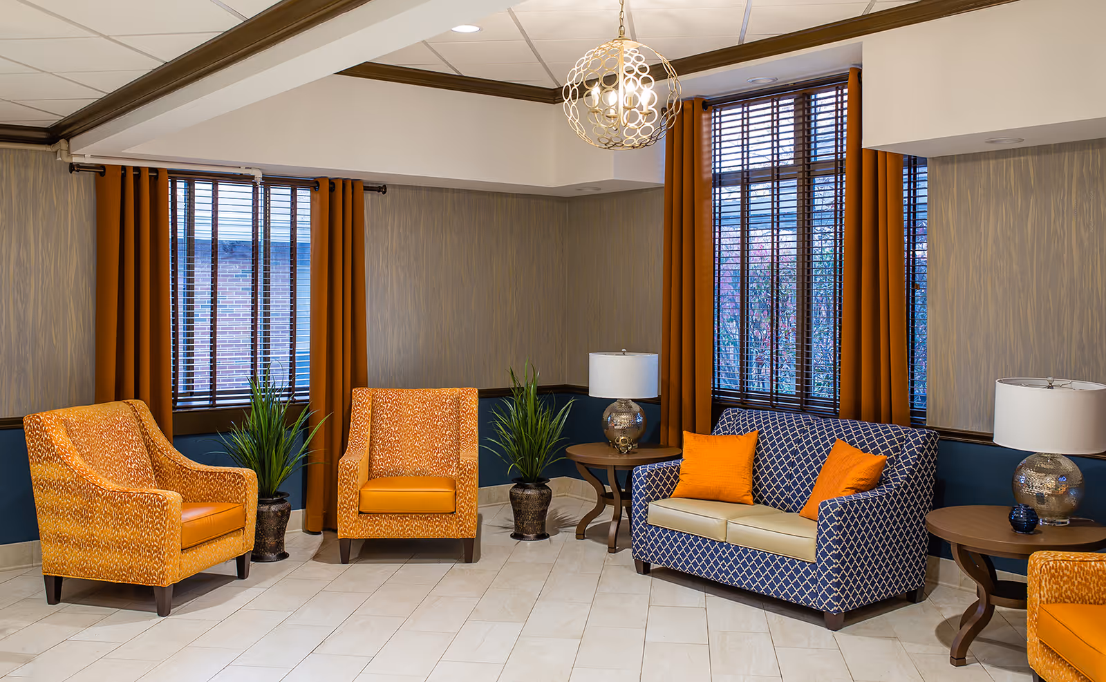 A lounge in a senior living facility with orange chairs, a blue patterned sofa, and lamps.
